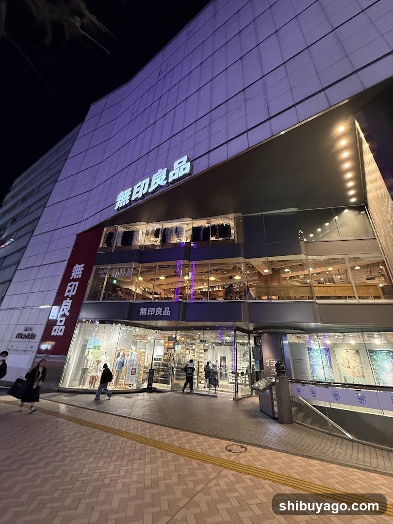 Night view of a Uniqlo store facade with illuminated signage displaying '無印良品' (Muji) and 'Uniqlo'. The building features large glass windows showing interior displays, mannequins, and clothing racks. Pedestrians walk on the sidewalk in front.