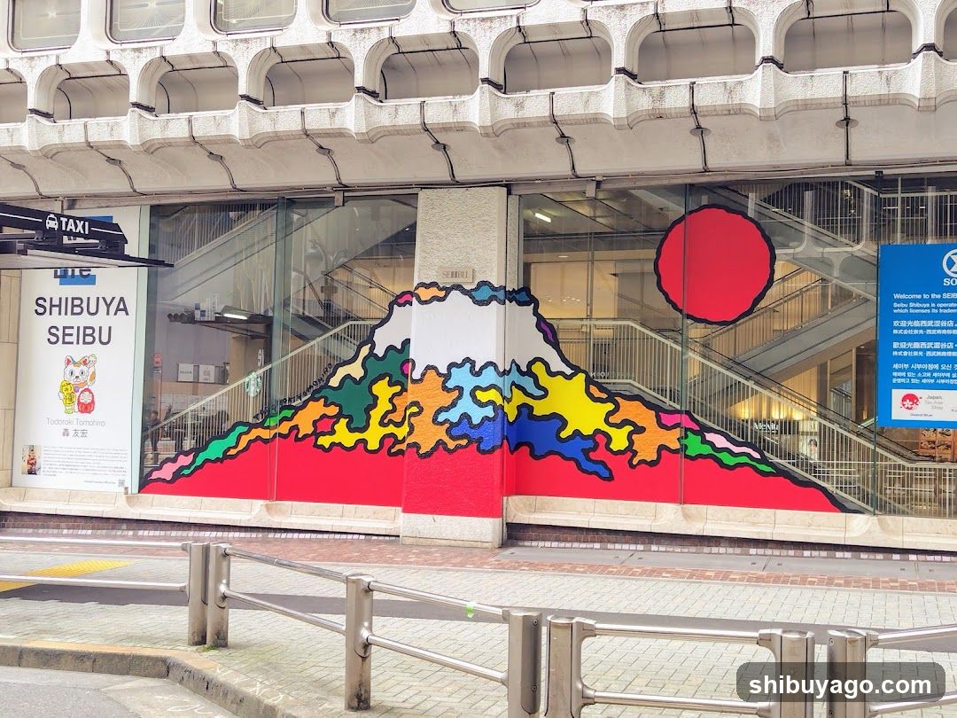The image shows the exterior of a Shibuya Seibu store, featuring a vibrant mural of Mount Fuji with colorful clouds and a red sun in the background. The building has large glass windows revealing an interior staircase. A taxi sign is visible on the left side, and there is a blue informational board on the right. The storefront includes signage indicating 'SHIBUYA SEIBU' along with Japanese characters.