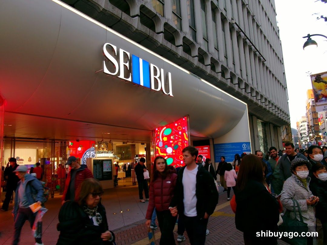 The image shows the exterior entrance of a Seibu department store, identifiable by its prominent illuminated signage on the building facade. The storefront features large glass windows and doors, with bright interior lighting visible inside. A colorful, abstract light installation is mounted near the entrance. Crowds of people are walking along the sidewalk in front of the store, suggesting a busy urban environment.