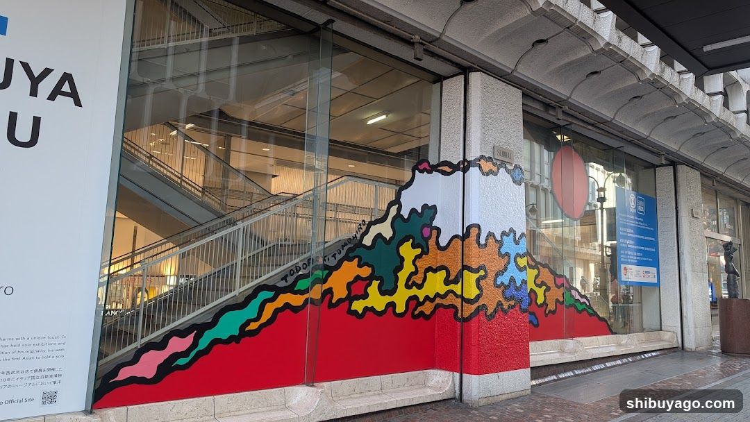 Exterior view of a modern building entrance featuring colorful graffiti art resembling Mount Fuji on the glass facade. Visible signage includes partial text 'UYA' and Japanese characters, along with informational plaques near the door.