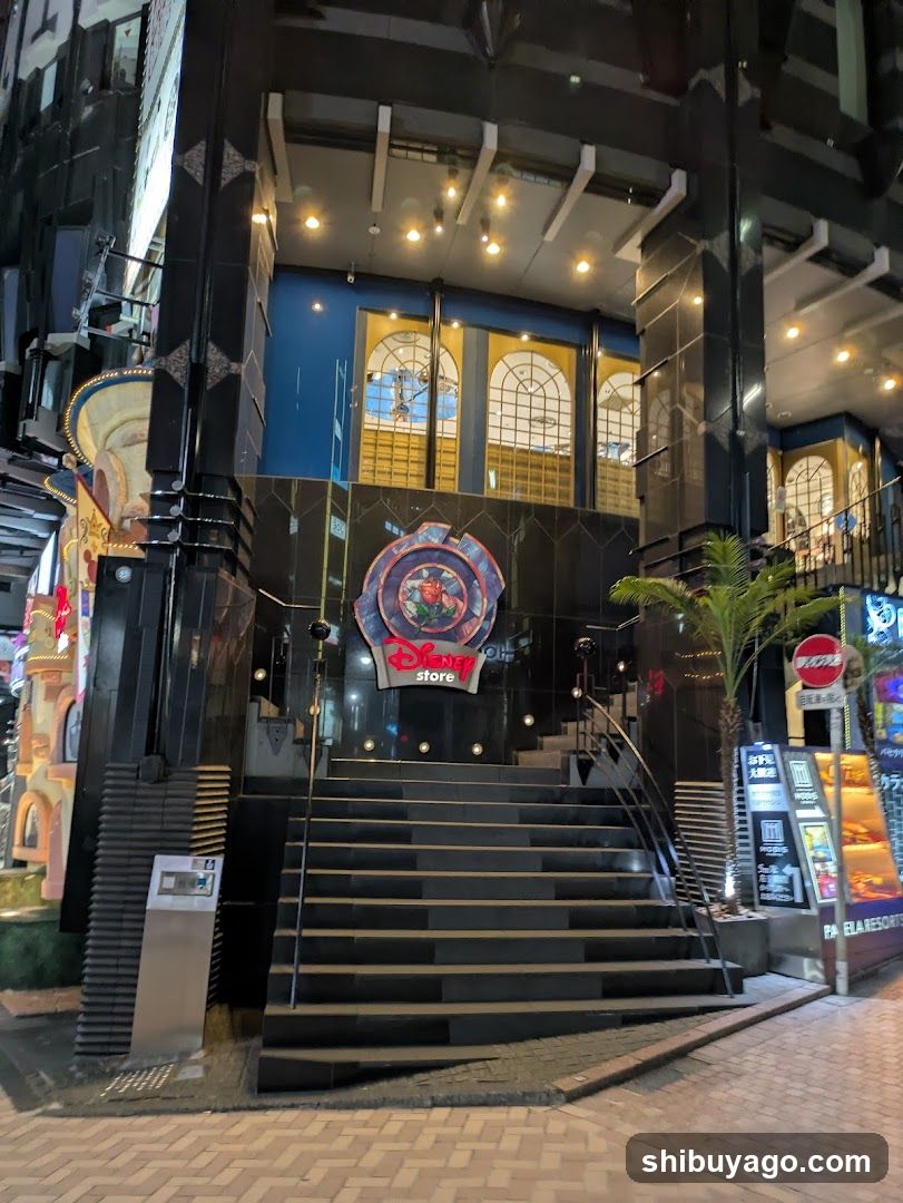 Night view of a modern building entrance featuring black tiled columns and steps leading up to glass doors. A circular Disney logo sign is mounted on the wall above the stairs. To the right, there are illuminated advertisements including a Coca-Cola sign and food menu boards displaying images of burgers and fries.