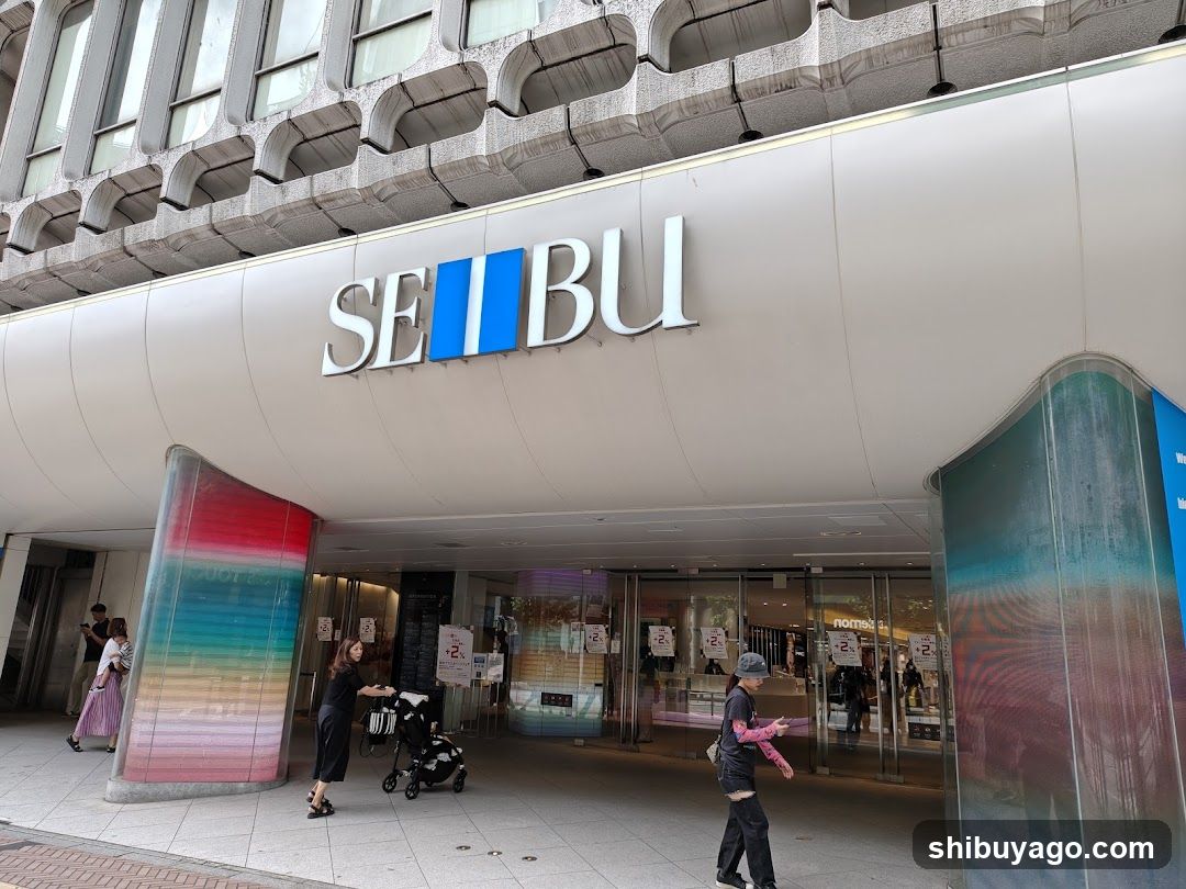 Photo of the physical exterior entrance/storefront facade of a Seibu department store, with visible signage on the building and people walking near the entrance.