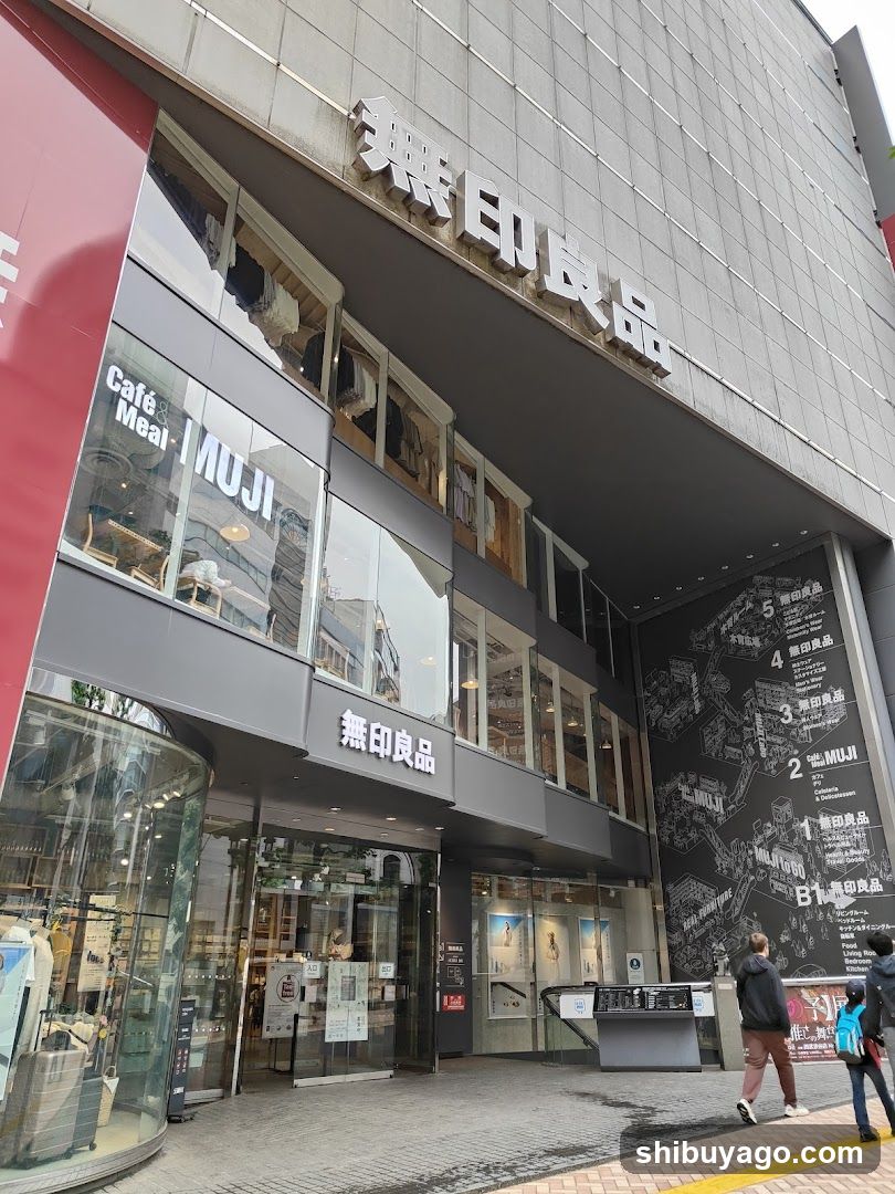 Photo of the physical exterior entrance and storefront facade of a MUJI store, featuring visible signage with Japanese characters '無印良品' and English 'MUJI', along with a 'Café Meal' sign above the entrance.