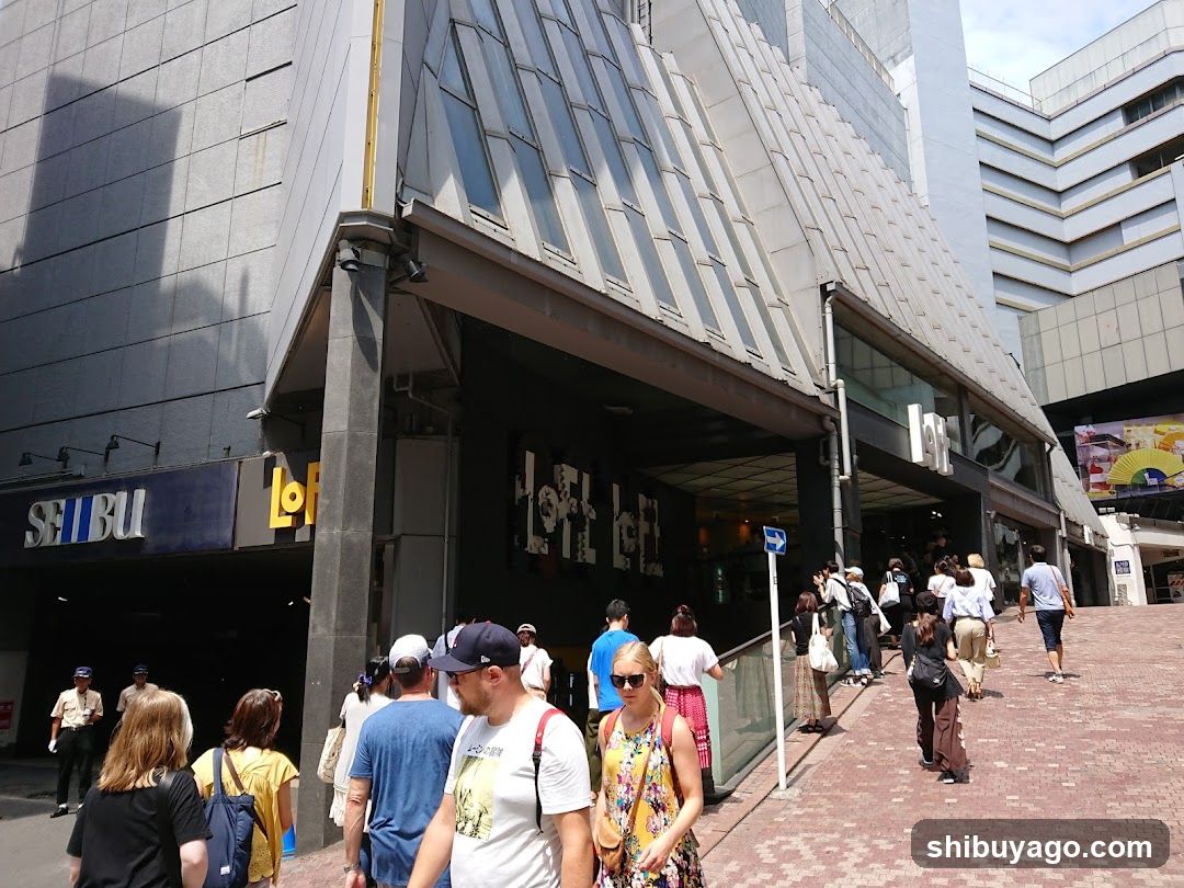 A busy urban street scene in front of a modern building with visible signage reading 'SHIBUYA' and 'LOFT'. People are walking on the sidewalk, some entering or exiting the store entrance. The architecture features angular glass panels and metal cladding.