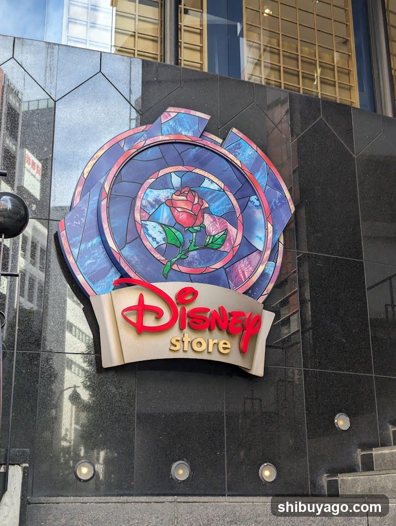 A photo of the physical exterior entrance/storefront facade featuring a prominent Disney Store sign mounted on a dark tiled building wall. The sign displays a stained-glass-style rose emblem above the red 'Disney' logo and gold 'store' text.