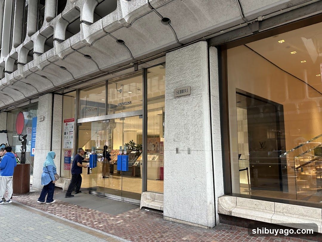 Photo of a modern building exterior featuring large glass windows and doors. Visible shop signage includes 'SEIBU' on a pillar. People are standing near the entrance, some holding blue bags. Interior displays include shelves and products visible through glass.