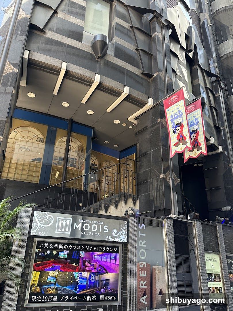 Exterior view of Modis Shibuya, a premium karaoke venue in Tokyo. The building features modern architecture with dark cladding and illuminated signage displaying 'MODIS SHIBUYA' and promotional banners for VIP rooms. Two Disney-themed flags hang near the entrance.