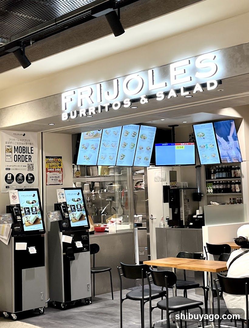 Storefront with illuminated signage reading FRIJOLES BURRITOS & SALAD. Digital menu boards display food items above the counter. Two self-order kiosks are positioned to the left. Seating area includes wooden tables and black chairs in front of the shop.