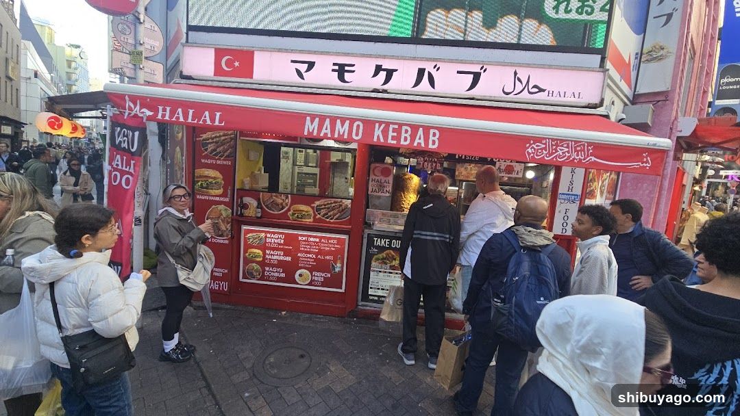 A bustling street scene featuring a red and white kebab shop with visible signage including 'HALAL' and 'MAMO KEBAB'. The storefront has large windows displaying food items, and customers are queuing outside.