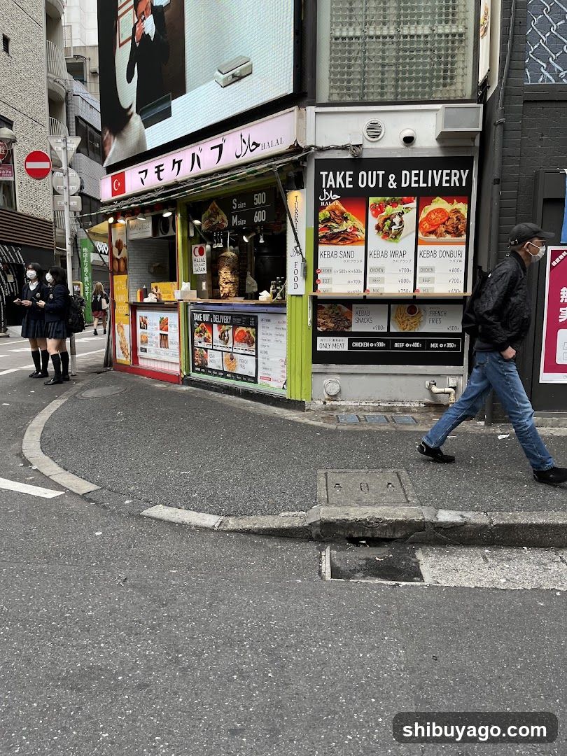 Street-level view of a kebab shop with visible signage and menu boards on the building facade. Two students in school uniforms stand near the entrance, while a pedestrian walks past. The shop features large windows displaying food items and takeout/delivery options.
