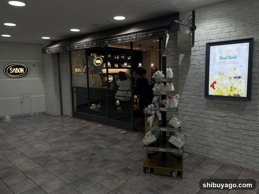 The image shows the exterior of a shop with a brick facade and large glass windows. A sign reading 'SABON' is visible on the left wall. Inside, shelves display various products. Outside, there is a rolling cart filled with packaged goods. An illuminated advertisement board is mounted on the right side of the building.