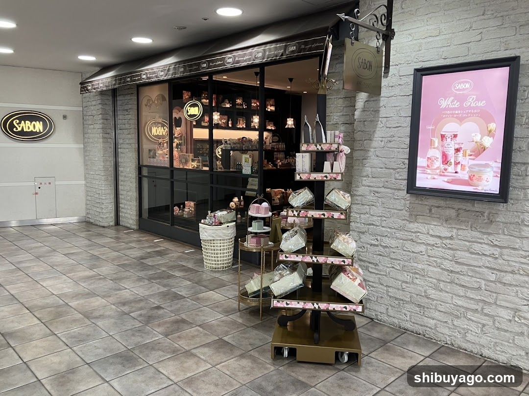 The image shows the exterior of a SABON store, featuring a brick facade with large glass windows displaying interior shelves filled with products. A prominent 'SABON' sign is mounted on the wall to the left. In front of the store, there are display racks showcasing various packaged items and a basket. An illuminated poster advertising 'White Rose' scented candles or similar products hangs on the right side of the building.
