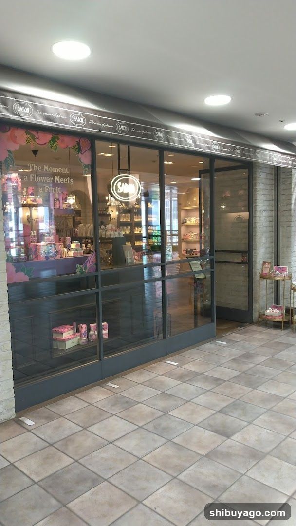 The image shows the exterior of a shop with large glass windows and doors, displaying various products inside. The sign above reads 'SAKURA' repeatedly along the top edge. Inside, there are shelves filled with items, including what appears to be floral arrangements or related merchandise. A neon sign with the word 'SAKURA' is visible through one of the windows. The floor outside is tiled, and additional small displays can be seen near the entrance.