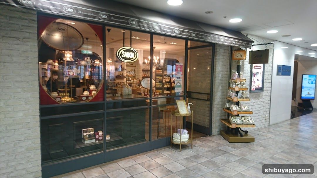 A shop with large glass windows and a brick facade, displaying various products inside. A hanging sign reads 'SAORI' above the entrance. Outside, there is a small display stand with packaged items.