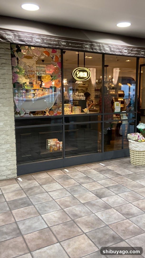 A shop front with large glass windows displaying products and a neon 'SABON' sign above the entrance. The left window features a floral advertisement, while the right shows interior shelves stocked with items.