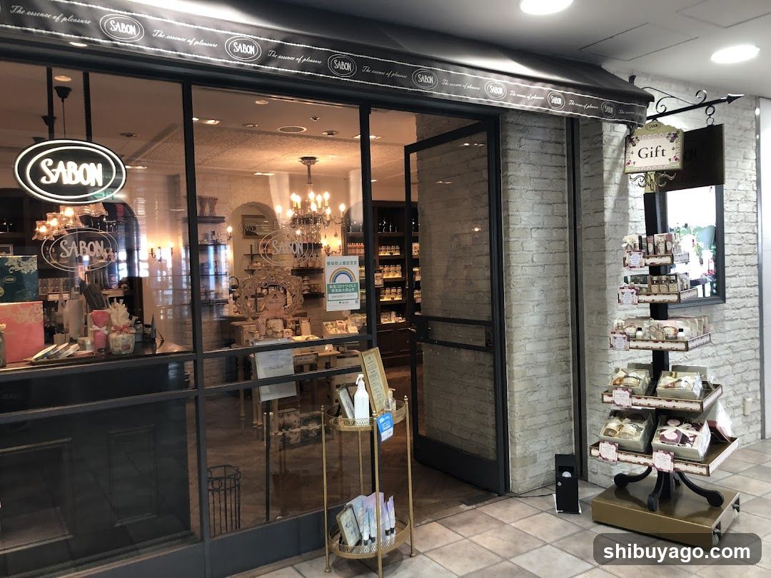 A shop front with large glass windows and doors, displaying the SABON logo prominently on the awning and window signage. Inside, there are shelves stocked with various products, chandeliers hanging from the ceiling, and a gift display stand outside.