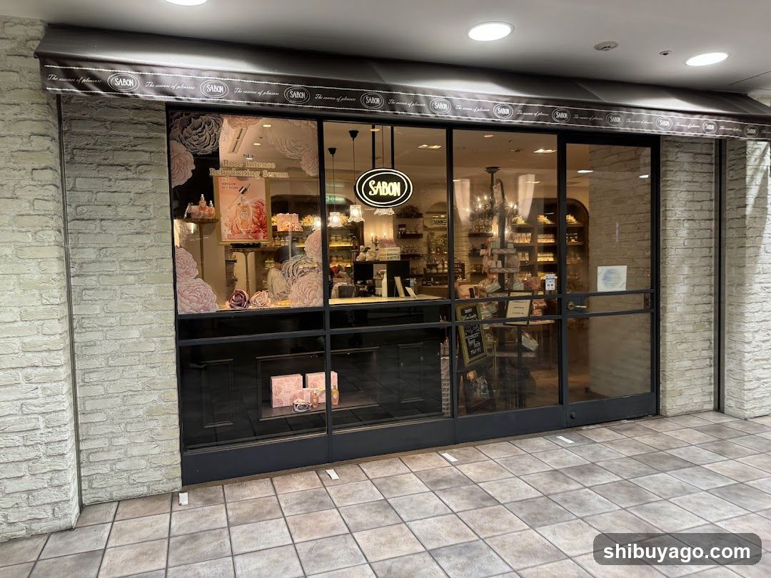 A shop front with a brick facade and large glass windows displaying interior shelves of products. A black awning with repeating 'SABON' logos runs above the entrance. Inside, hanging lights illuminate the space, and a circular neon sign reading 'SABON' is visible near the center. The floor outside is tiled, and promotional posters are affixed to the window panes.