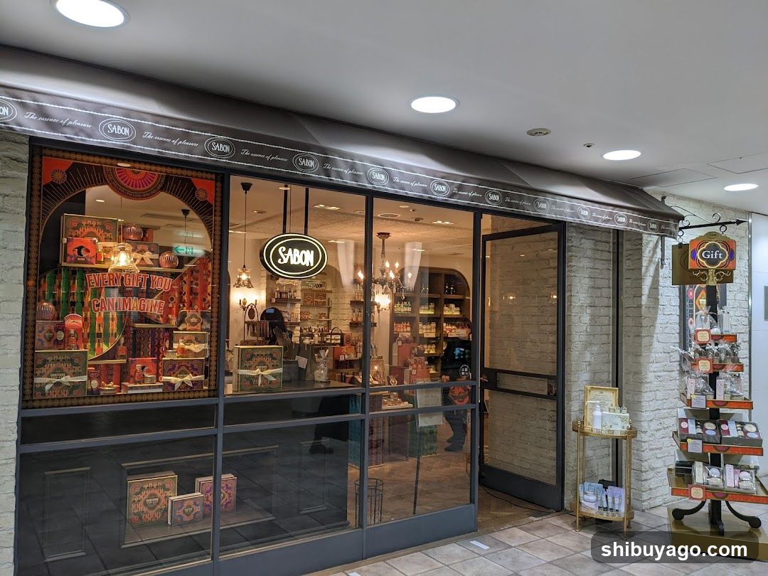 A shop front with a large glass facade and an awning displaying the SABON logo repeatedly. The entrance is flanked by brick walls, and there are promotional displays outside. Inside, shelves stocked with products are visible through the windows.