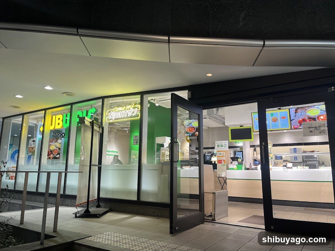 Night view of a Subway restaurant entrance with illuminated signage reading 'SUBWAY' and 'Come on NIGHT'. Glass doors are open, revealing interior counter area with menu boards displaying food items above the service window.