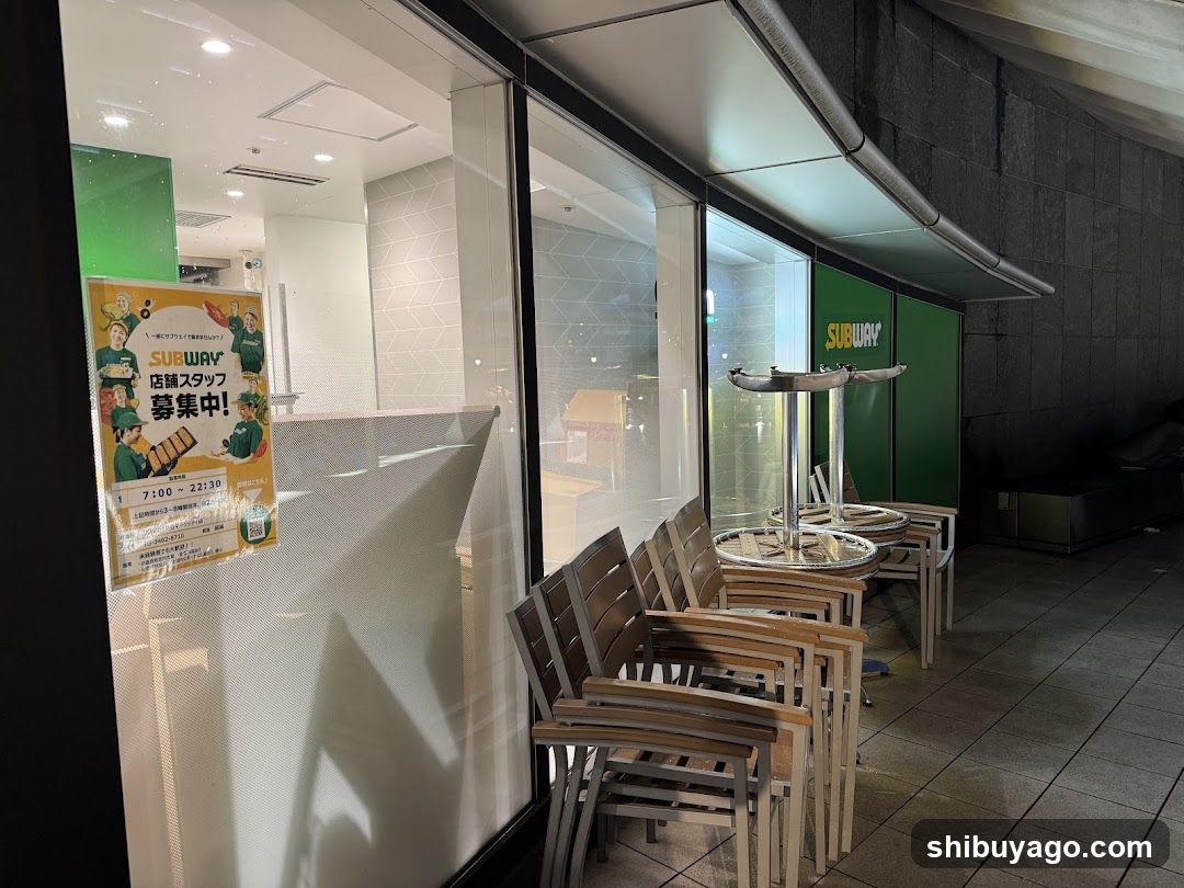 Photo of physical exterior entrance/storefront facade with visible shop signage. A Subway poster is displayed on the glass window, advertising staff recruitment. Outdoor seating area with stacked chairs and tables is visible outside.