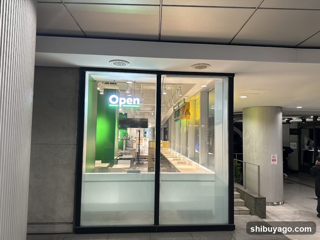 A modern storefront featuring large glass windows and doors, displaying an illuminated 'Open' sign inside. The interior shows a clean, minimalist design with green accents and display counters.