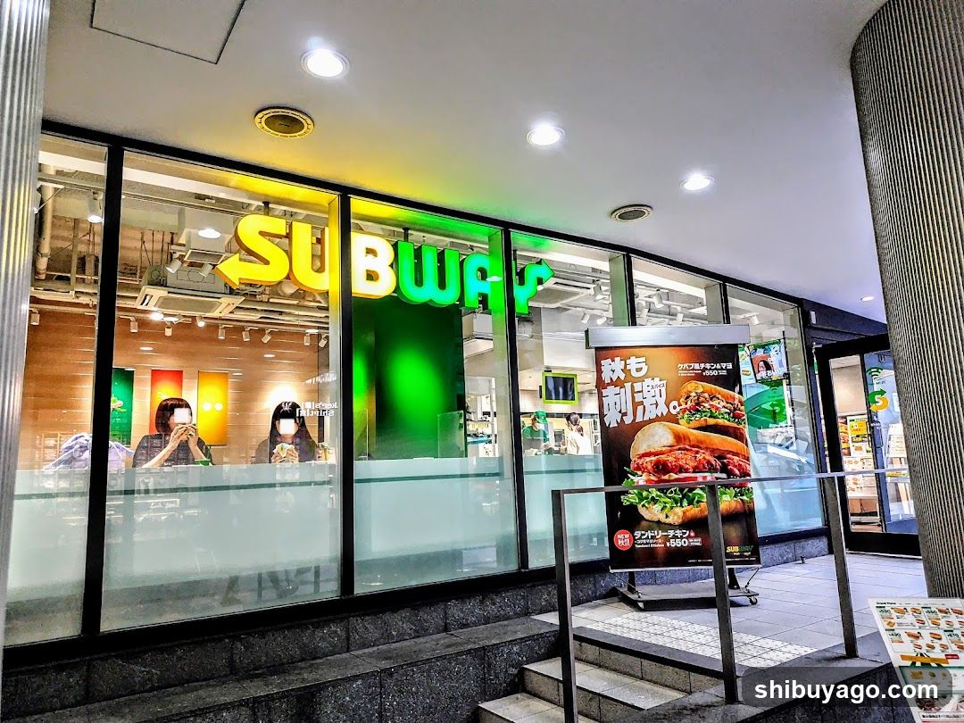 A Subway restaurant exterior with illuminated signage and a promotional poster featuring autumn-themed sandwiches.