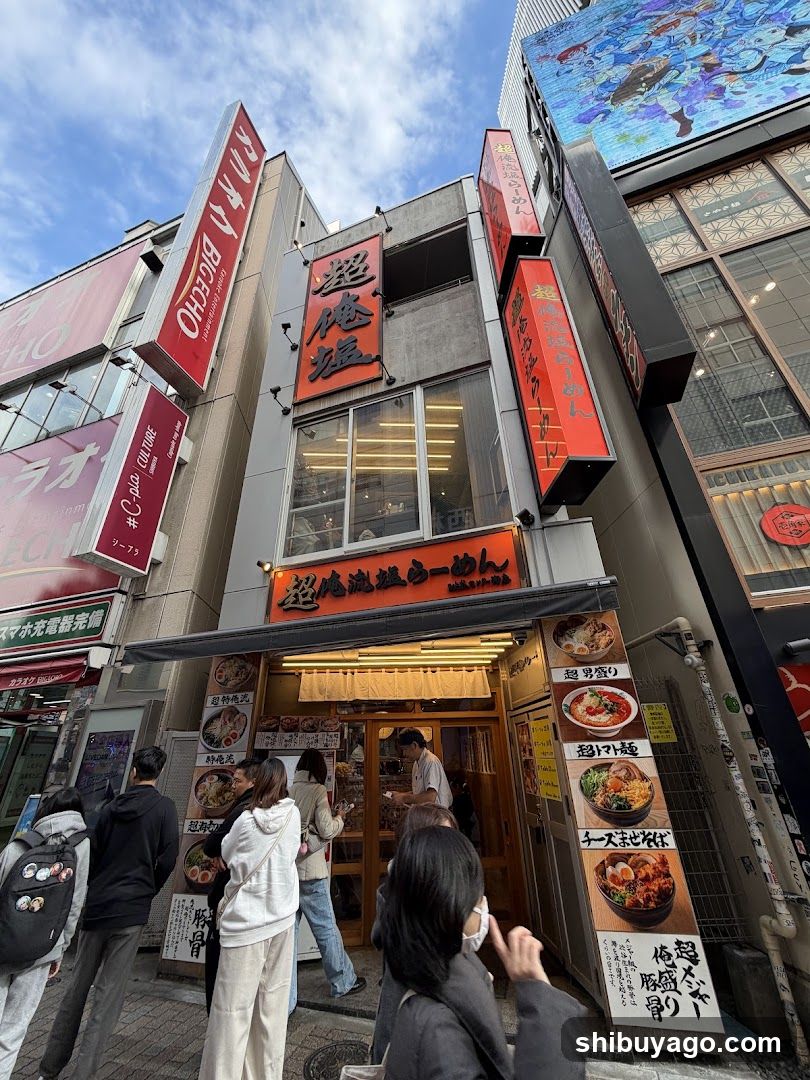 A street-level view of a ramen shop with prominent red and orange signage featuring Japanese characters. The entrance is open, revealing the interior where staff are visible. Several customers stand outside near the entrance. Vertical banners hang on either side of the building facade.