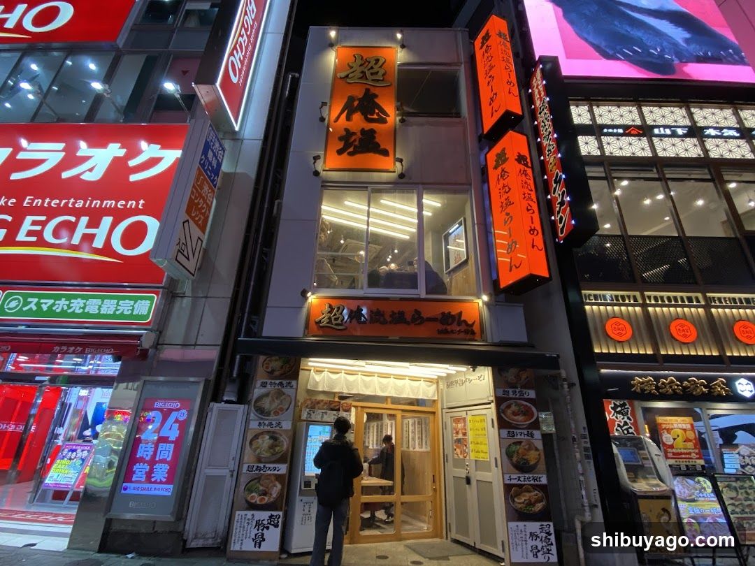A nighttime street scene featuring multiple storefronts with illuminated signage. The central building has vertical orange signs reading '超俺流' (Chou Ore Ryu) and a horizontal sign below the entrance. To the left, a red sign displays 'ECHO' and 'Entertainment'. On the right, another establishment shows circular neon signs and glass windows displaying food items inside. A person stands near the entrance of the central shop.