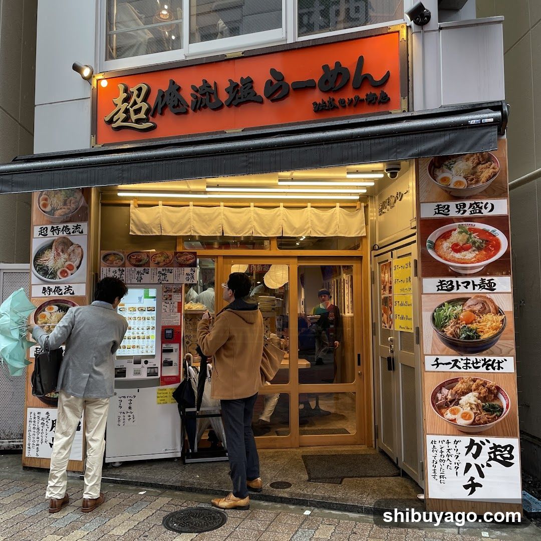 A photo of the physical exterior entrance and storefront facade of a ramen shop in Japan. The orange sign above reads '超絶流塩ラーメン' (Choketsu Ryuen Ramen). Two customers are standing at the entrance, one holding an umbrella. A menu board is visible on the right side of the entrance.