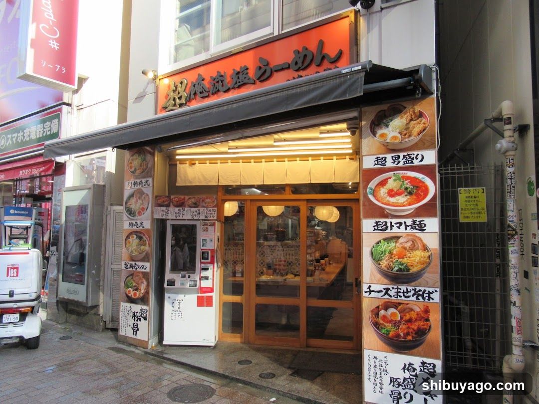 A street-level view of a ramen shop with an orange sign reading '超味流麺ラーメン'. The entrance features glass doors and windows, displaying interior seating. A vertical menu board on the right lists dishes like '超男盛り', '超いなり麺', 'チーズまめ丼', and '超盛り豚骨'. Adjacent signs advertise '特製豚骨' and '豚骨'. A white vending machine stands to the left.