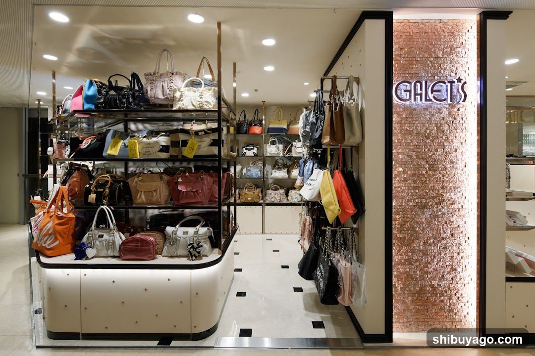 Interior view of a boutique store featuring shelves filled with handbags and accessories. A prominent illuminated sign reading 'GALET'S' is mounted on a textured brick wall to the right. The display includes hanging bags, stacked handbags in various colors (orange, blue, black, beige), and a white countertop with additional items. Lighting fixtures are visible above, illuminating the merchandise.