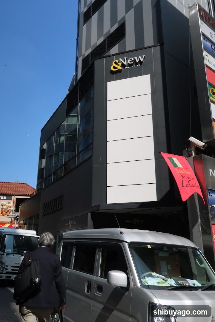 A modern building facade featuring a prominent "&New" sign on the upper section. Below, there is a red flag with Italian text and an image of pasta. A silver van is parked in front, partially obscuring the lower part of the building. The sky is clear blue.