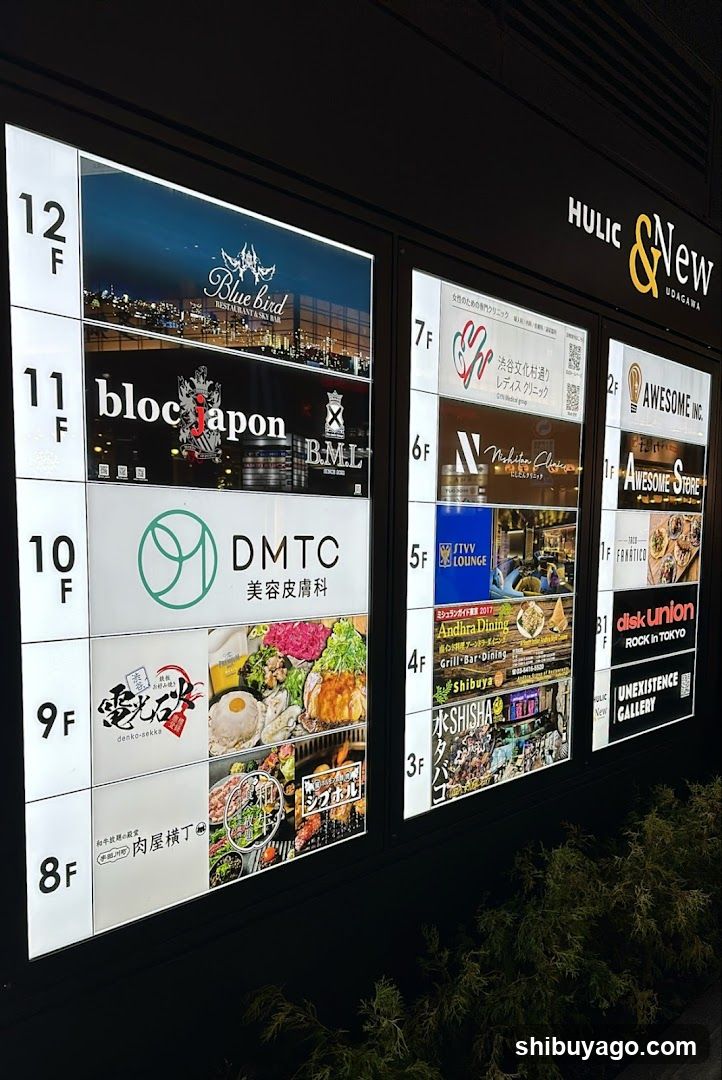 Vertical illuminated directory board on building exterior showing floor levels from 8F to 12F with corresponding tenant signage including Bluebird, blocjapon, DMTC, and various restaurants like Awesomeness Inc., disk union, and Shishamo.