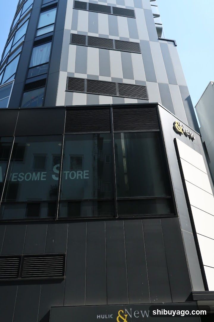The image shows the exterior of a modern building featuring a glass storefront with visible signage reading 'AWESOME STORE' on the window. The facade includes geometric patterns and ventilation grilles, with additional branding at the bottom displaying 'HULIC & New'. No menu or interior details are visible.