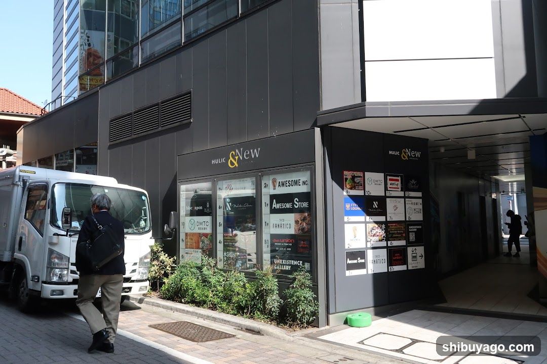 Photo of the exterior entrance and storefront facade featuring visible signage including 'HOLIC & NEW' above glass windows displaying various posters and advertisements. A white truck is parked nearby, with a pedestrian walking past.