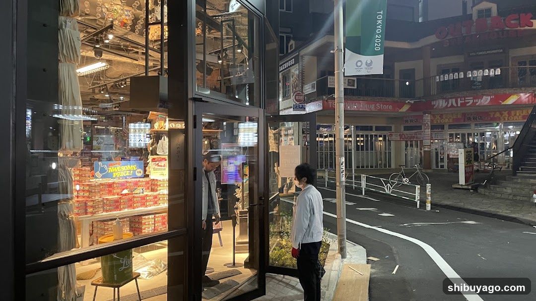 Night view of a shop entrance with glass doors and illuminated interior displaying stacked products. A person stands outside near the door, another inside. Background shows street signage including 'OUTBACK STEAKHOUSE' and Japanese text banners.
