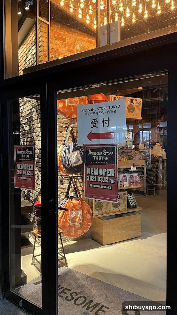 The photo shows the storefront of Awesome Store Tokyo, featuring glass doors with signage indicating a new opening date of March 12, 2021. Inside, there are shelves stocked with various products and decorative elements like hanging lights and brick walls.