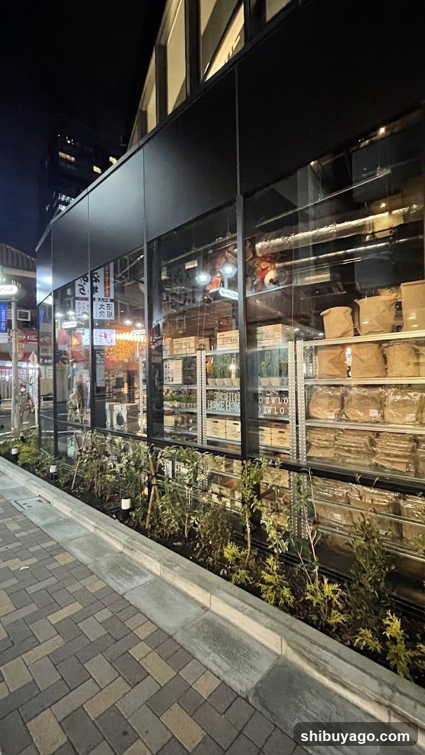 A modern shop front at night featuring large glass windows displaying shelves stocked with packaged goods, likely food items. The exterior includes a planter box with small plants along the sidewalk. Signage is visible on the building facade.