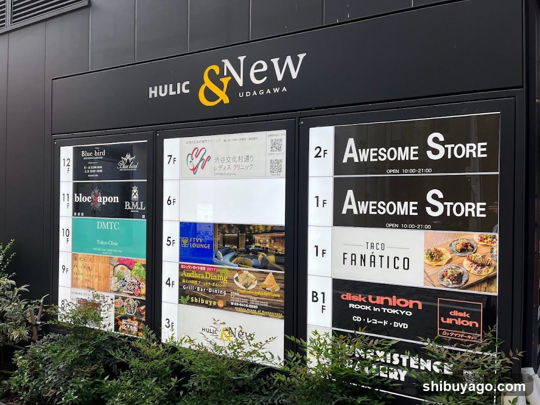 Exterior storefront of HULIC & New Udagawa, a multi-level shopping complex with visible signage and floor directory panels displaying various shops like Awesome Store, Taco Fanático, and Disk Union.