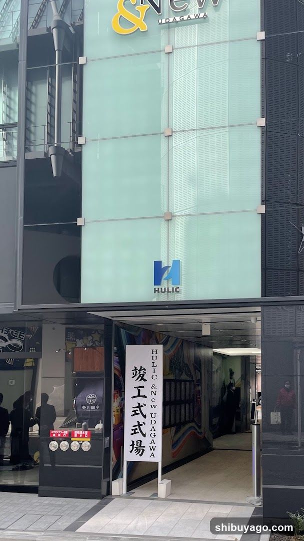 Exterior view of a modern building entrance featuring glass facade with 'HULIC' and '& NEW HUGAWA' signage. A vertical banner displays Japanese characters for 'Grand Opening'. The storefront includes illuminated signs, vending machines, and visible interior walkway.