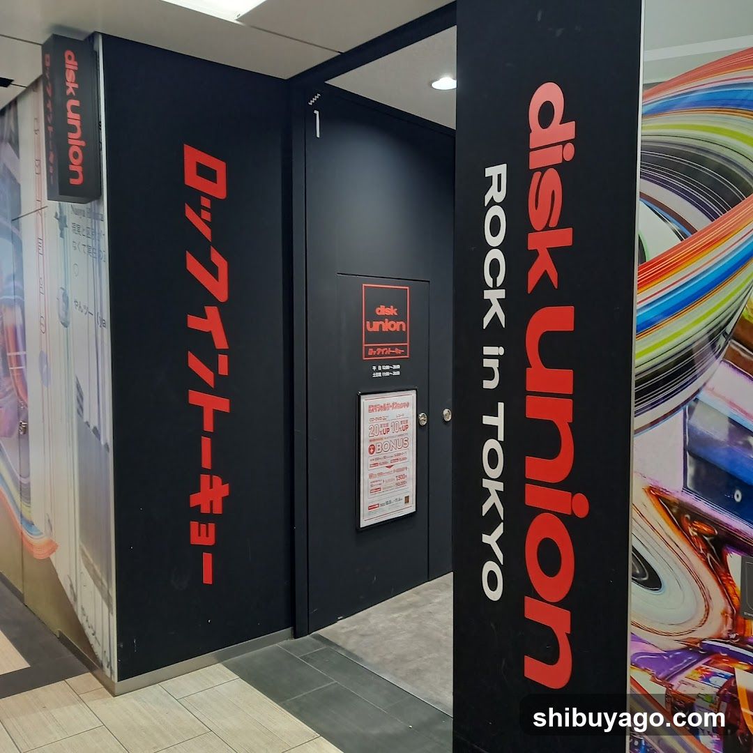 Photo of the physical storefront entrance to disk union in Tokyo, featuring black walls with red and white signage displaying 'disk union' and Japanese text. A poster board is mounted on the door.