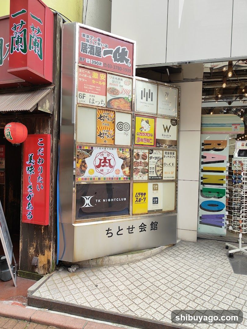 A street-level view of a building facade featuring multiple signs. On the left, a red sign with green Japanese characters is mounted above an entrance with a hanging red lantern. To its right stands a tall metal kiosk displaying numerous advertisements for various businesses including 'Ikkoku', 'Hatcho', 'TK Nightclub', and others. Below these ads, the name 'ちとせ会館' (Chitose Kaikan) is written in large black characters on a silver panel. Further to the right, part of another shop's interior is visible through glass doors, along with a rack holding colorful foam letters.