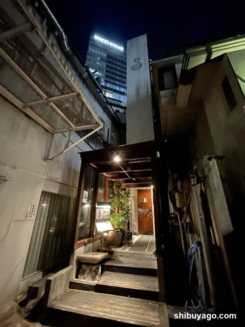Night view of a narrow alleyway entrance with illuminated storefront and modern building in background