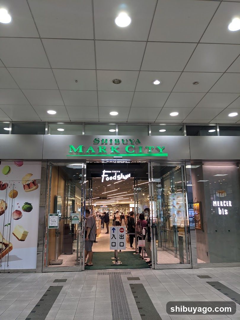 Photo of the physical exterior entrance storefront facade with visible signage 'SHIBUYA MARK CITY' in green letters above glass doors. A poster on the left shows food items like sandwiches and fruit. Inside, people are walking through the mall area.