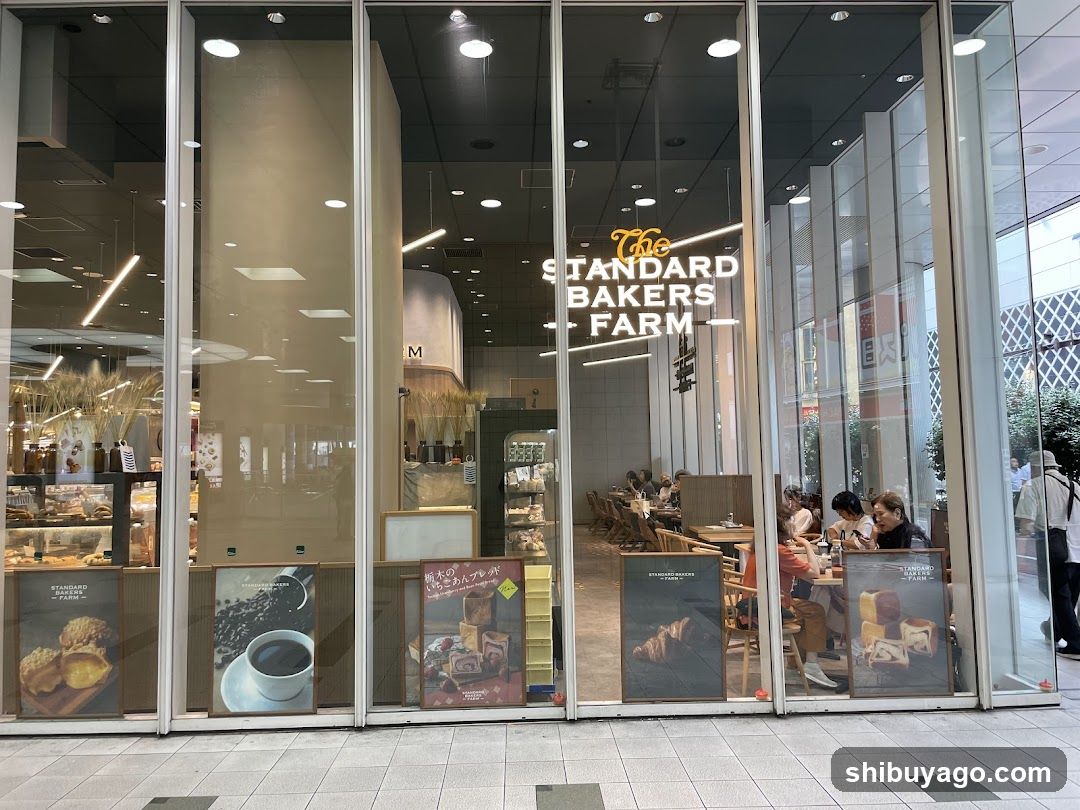 A modern storefront with large glass windows displaying interior seating and baked goods. Visible signage reads 'The STANDARD BAKERS FARM'. Posters at the bottom advertise pastries, coffee, and seasonal items.