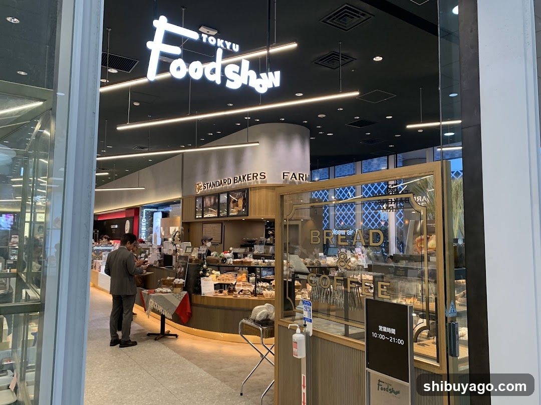 The image shows the entrance of a Tokyu Foodshow store, featuring illuminated signage with 'TOKYU Foodshow' prominently displayed above. Inside, there is a bread and coffee section labeled 'BREAD & COFFEE', along with other food items visible on display counters. A person is seen standing near the counter, possibly ordering or browsing.