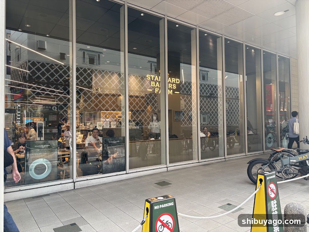 A modern storefront with large glass windows displaying interior seating and staff. Visible signage includes 'STANDARD BAKERS' on the window. Outside, yellow 'NO PARKING' signs are placed near a parked motorcycle. Inside, customers sit at tables while staff work behind counters.