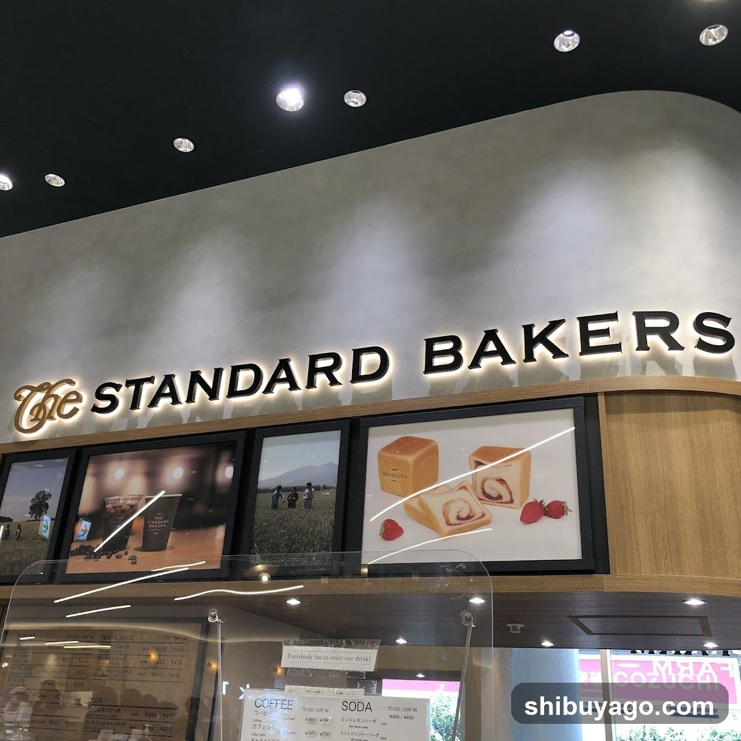 A modern bakery storefront with illuminated signage reading 'The STANDARD BAKERS'. Below the sign, there are four framed promotional images showcasing baked goods like bread and pastries. The entrance features glass doors with reflections of interior elements. A small menu board is visible inside through the glass.