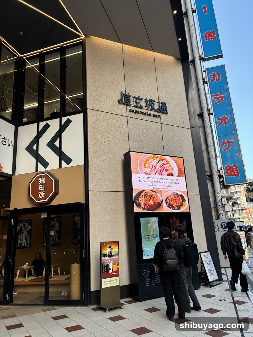Exterior view of a shop entrance with signage reading '道玄坂通' and 'dogenzaka street'. A digital display shows food images, including ramen dishes. People are standing near the entrance.