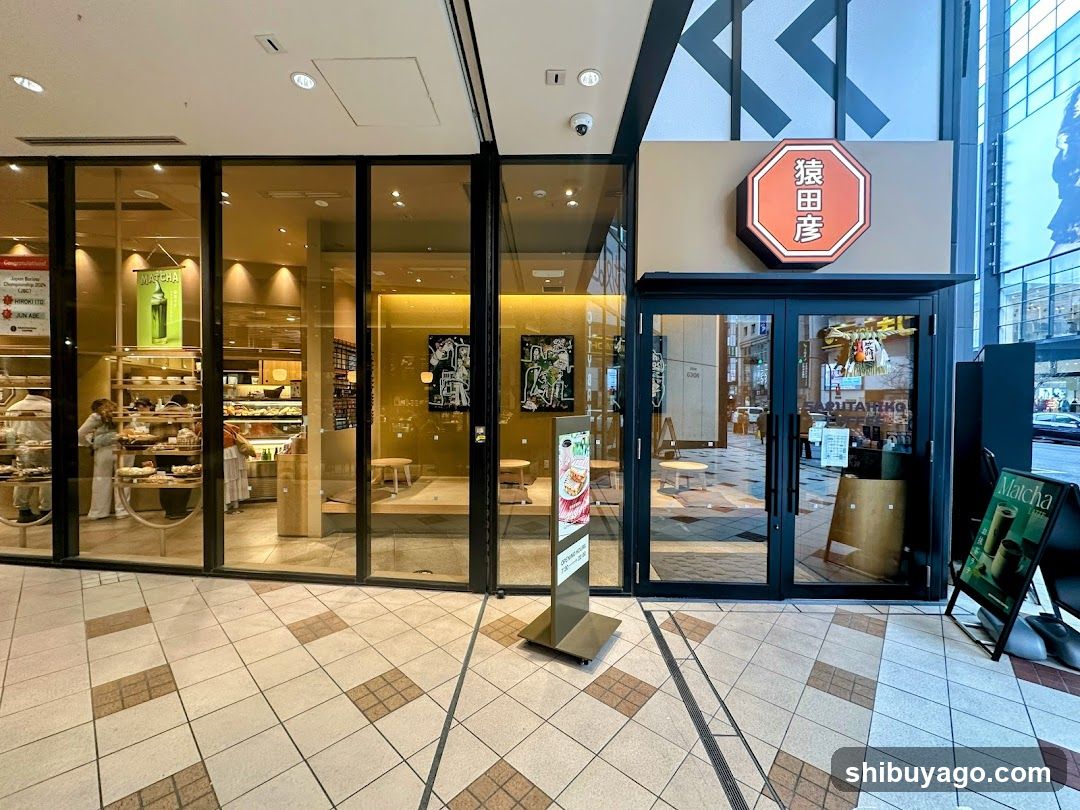 A modern storefront with large glass windows and doors, featuring a prominent red octagonal sign with Japanese characters above the entrance. Inside, shelves display various products, and a person is visible near the counter.